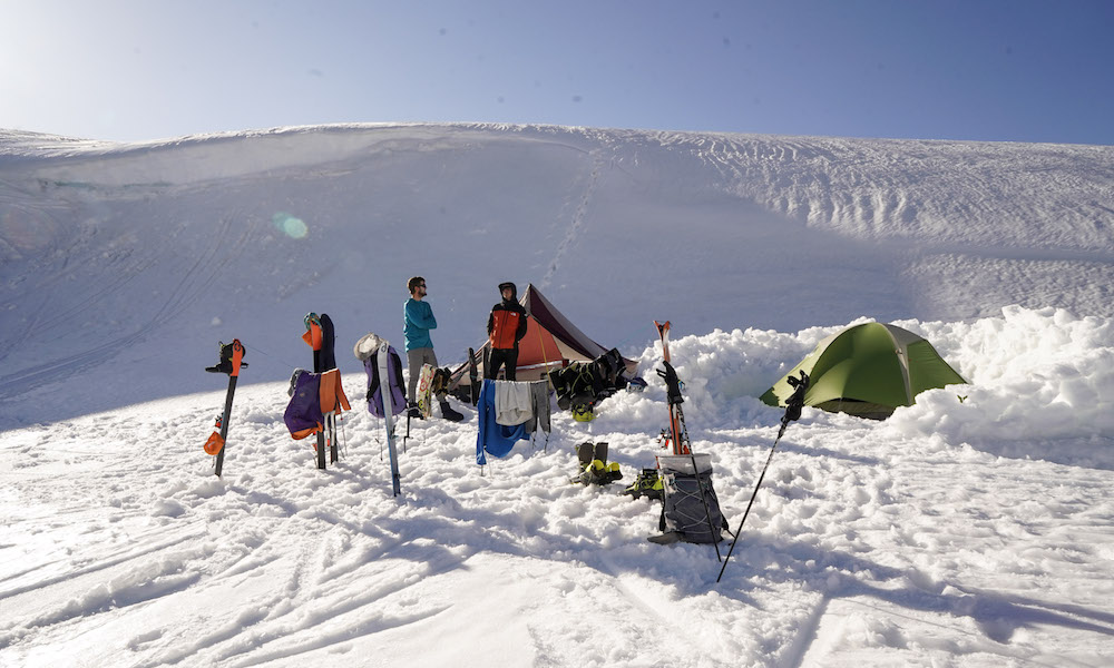 A photo of our final basecamp. Gear is drying on strung-out lines, water is boiling in the cooktent, the bootpack is set for cornice sledding, and a snow wall helps limit our exposure to the wind. An open stream is 200' away from the camp, and the hill behind camp helps protect us from larger gusts of wind sweeping from across the lake.