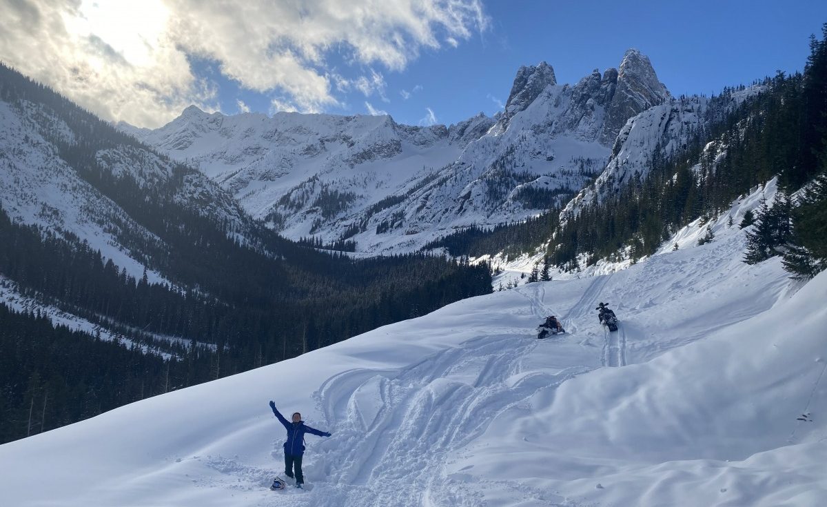 Some of the old avalanche debris visible on the road just past the snowmobiles, just a little below the top of Washington Pass. The spires visible and gorgeous, Kristen looking stoked to be able to finally get out in this area.